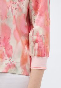 Μανίκι ροζ και πορτοκαλί πουκάμισου με σχέδιο γεωμετρικών γραμμών και ριμπ μανσέτα, φορεμένο πάνω από λευκό παντελόνι.