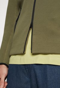 Olive green zip-up jacket with black zippers and hem, layered over a yellow shirt, paired with blue herringbone-patterned pants.