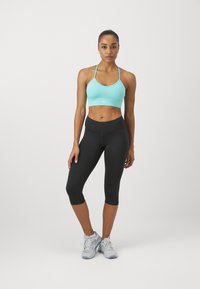 Soutien-gorge sport azzurro con spalline sottili, abbinato a leggings neri a tre quarti. Il modello indossa scarpe da ginnastica grigie e blu. Design semplice e funzionale.