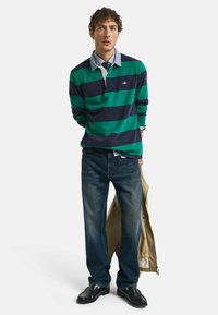 Poloshirt met lange mouwen in marineblauw en groene horizontale strepen, met een witte kraag en logo. Gepaard met donkere spijkerbroek en zwarte schoenen.