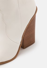 Bílá kotníková bota z kůže s hnědým blokovým podpatkem a detailním prošíváním na bílém pozadí.