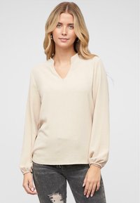 Blusa beige a maniche lunghe con scollatura a V, polsini arricciati e vestibilità rilassata, realizzata in tessuto leggero con una consistenza morbida.