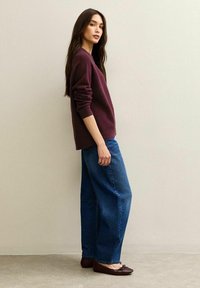 Maglione oversize bordeaux a maglia, jeans blu larghi e ballerine scure. Modella in posa di lato contro uno sfondo neutro. Design minimalista.