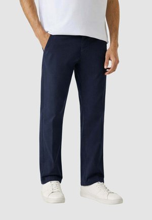 Uomo che indossa pantaloni dritti blu navy, scarpe da ginnastica bianche e una camicia bianca con le mani parzialmente nelle tasche, in piedi contro uno sfondo semplice.