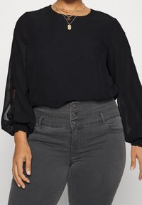 Blouse noire avec manches bouffantes transparentes et encolure ronde, associée à un jean gris taille haute avec une fermeture à boutons sur le devant.