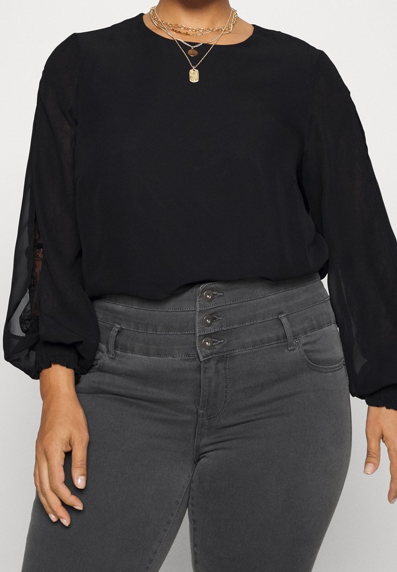 Blouse noire avec manches bouffantes transparentes et encolure ronde, associée à un jean gris taille haute avec une fermeture à boutons sur le devant.