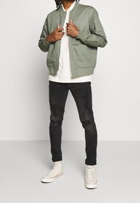 Zielona bomberka z prążkowanymi mankietami, biała koszulka pod spodem, czarne dżinsy z przetarciami i białe trampki na jednolitym tle.