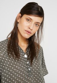 Chemise à motifs en vert foncé avec des designs géométriques, à manches courtes et avec des boutons. Tissu lisse et léger avec une coupe décontractée.