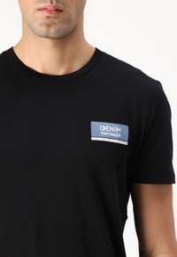 Sort t-shirt med korte ærmer, der har et blåt og hvidt logo-patch på venstre brystområde, lavet af blødt og glat stof.
