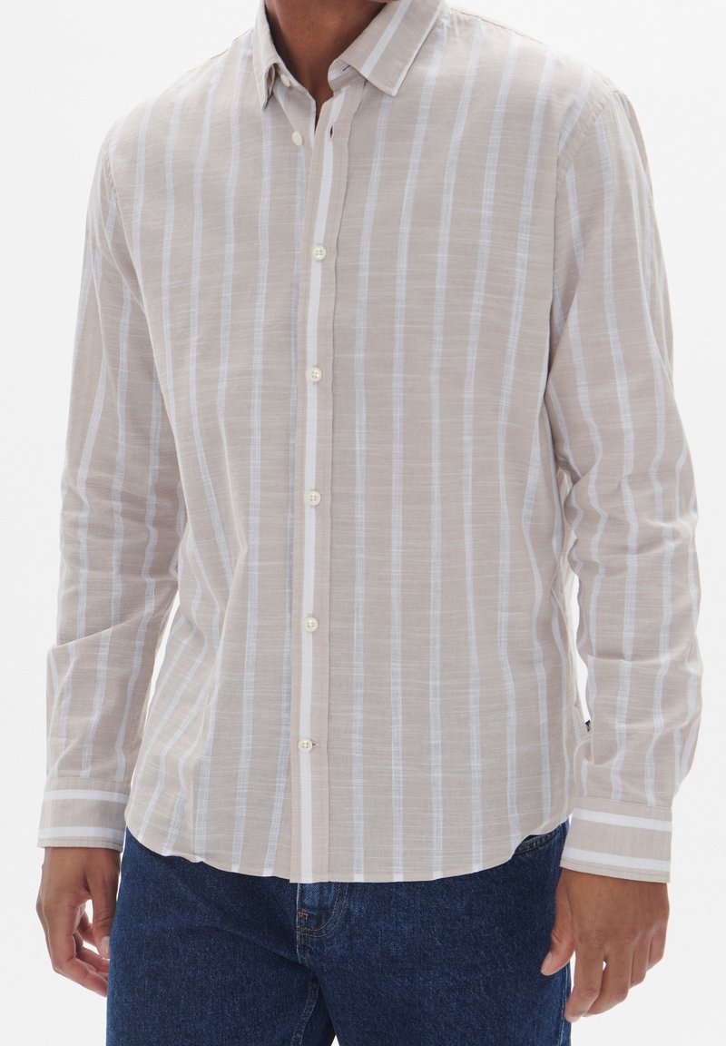 Homme portant une chemise à manches longues beige et blanche à rayures verticales, avec un jean bleu, debout les bras relâchés le long du corps.
