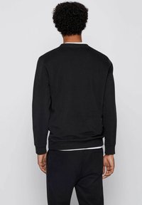 Hombre con cabello rizado, vistiendo un sudadera negra de manga larga y pantalones negros, de pie frente a un fondo liso de color gris claro.