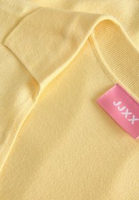 Gilet tricoté jaune pâle avec un poignet replié et une étiquette en tissu rose portant la mention "JJXX" cousue à l'intérieur du col.