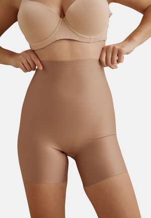 Femme portant un soutien-gorge beige et un shorty gainant taille haute, ajustant sa taille sur un fond blanc uni.