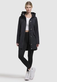 Black waterproof jacket with a hood, zippered front, and snap buttons; worn over a white crop top and black leggings with graphic accents.