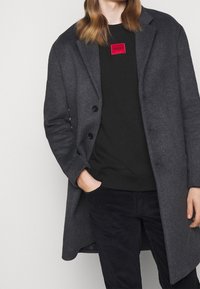 Cappotto di lana grigio scuro con design a un petto, bottoni neri e una texture morbida, abbinato a una felpa nera con logo rosso.