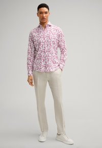 Weißes Hemd mit pinkem Blumenmuster, mit Knöpfen, langen Ärmeln; kombiniert mit hellbeigen Leinenhosen und weißen Sneakers.