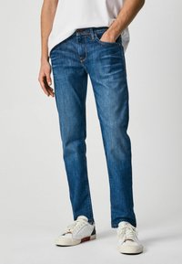 Vaqueros de mezclilla azul con corte recto, diseño de cinco bolsillos y un desgaste sutil. Combinados con zapatillas blancas y una camiseta blanca sencilla.