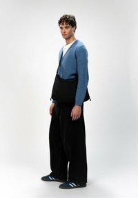 Blauer gestrickter Cardigan über einem weißen Hemd, kombiniert mit weiten schwarzen Hosen und blauen Sneakers. Eine schwarze Umhängetasche ruht auf der Hüfte.