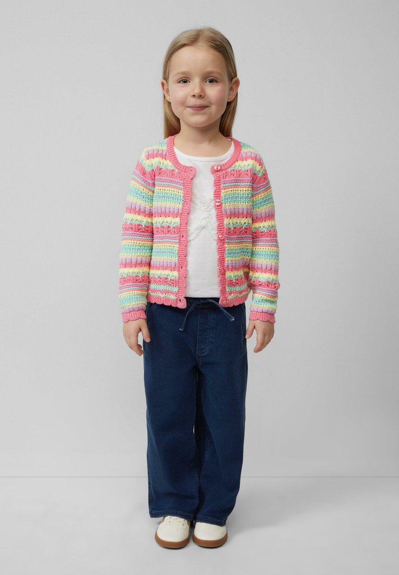 Mehrfarbige Strickjacke mit pinken, blauen und grünen Streifen, kombiniert mit einem weißen Hemd und dunkelblauen weiten Hosen, getragen mit weißen Schuhen.