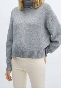 Suéter gris de punto con cuello alto, dobladillo acanalado y corte holgado, combinado con pantalones ajustados de un beige claro. Textura suave visible.
