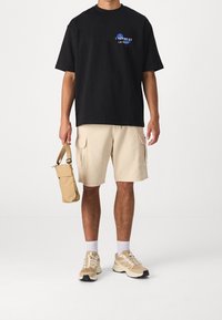 Černé tričko s krátkým rukávem a modrým grafickým textem, béžové cargo kraťasy, béžové tenisky a malá béžová taška.