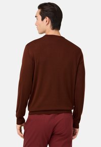 Rjav pleten pulover z dolgimi rokavi, okroglo izrezom in rebrastimi manšetami, v kombinaciji z burgundskimi hlačami, viden od zadaj.