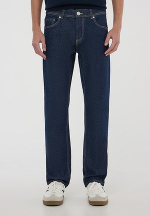 Jeans in denim blu scuro con taglio dritto, cinque tasche, cuciture gialle e chiusura con bottone. Indossati con scarpe da ginnastica bianche e grigie.