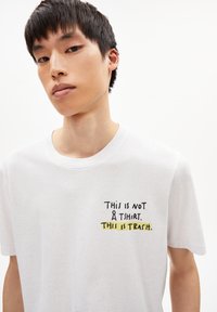 Witte katoenen T-shirt met een ronde hals. Voorzien van zwarte en gele tekst: "DIT IS GEEN T-SHIRT. DIT IS AFVAL." Minimalistisch ontwerp.