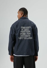 Marineblauw geruit overhemd met lange mouwen en geborduurde tekst op de achterkant. Heeft een losse pasvorm en knoopmansetten. Gemaakt van een lichtgewicht stof.