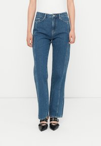 Jeans de mezclilla azul con corte recto, diseño de cinco bolsillos y costuras laterales en contraste. Combinados con zapatos negros de punta. 