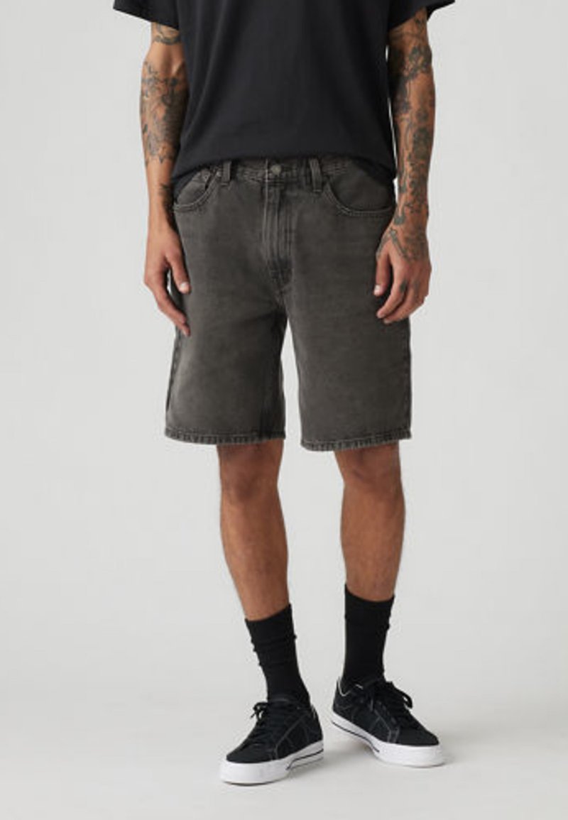 Person iført falmede sorte denimshorts, sorte crew-strømper, sorte sneakers og en sort t-shirt, stående med hænderne afslappet langs siderne.