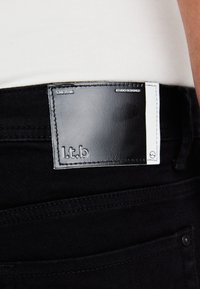 Svarta jeans i denim med en läderetikett sydd vid midjan, med präglingstekst "l.t.b". Etiketten har en vit kant med tryckta detaljer.