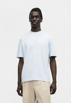 Ung mand med mørk hud og korte dreadlocks iført en lyseblå oversized T-shirt og beige bukser, stående mod en ensfarvet baggrund.