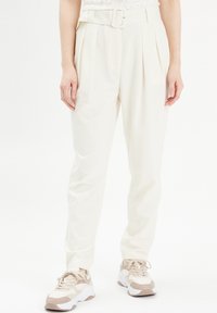 Pantalons taille haute de couleur crème avec une ceinture, dotés de plis et d'une texture lisse, associés à des baskets de teinte claire.