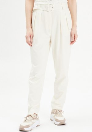 Cream-colored high-waisted trousers with a belt, featuring pleats and a smooth texture, paired with light-toned sneakers.