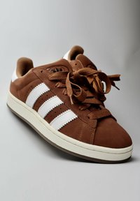 Brown suede sneaker with white stripes, round toe, textured surface, and flat brown laces. Features a rubber sole and perforations for breathability.