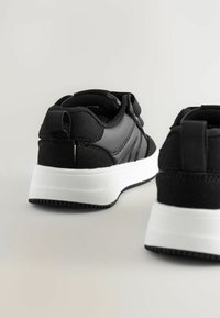Baskets de sport noires avec une tige texturée, dotées d'une semelle blanche, d'une languette et d'éléments de design superposés sur les côtés.