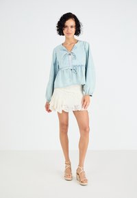 Ljusblå denimblus med knytframs, långa ärmar och samlad midja, tillsammans med en vit, fransad lace minikjol och sandaler med kilklack i tan.