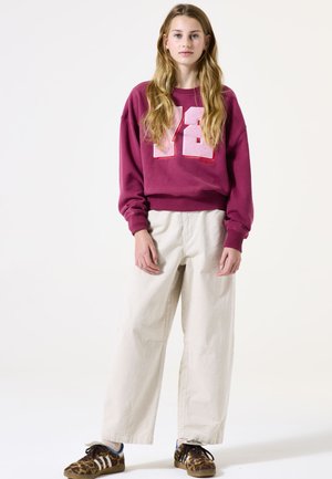 Sweatshirt surdimensionné bordeaux avec impression texturée blanche '82', associé à un pantalon large crème et des chaussures à imprimé léopard brun.