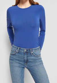 Top de mangas largas azul hecho de una tela suave y elástica; presenta un escote redondeado y se ajusta al cuerpo, combinado con jeans de mezclilla.