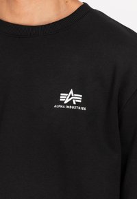 Svart hettegenser med ribbet hals, med et hvitt logo med et 'A' og tre striper på venstre bryst. Mykt bomullsmateriale.