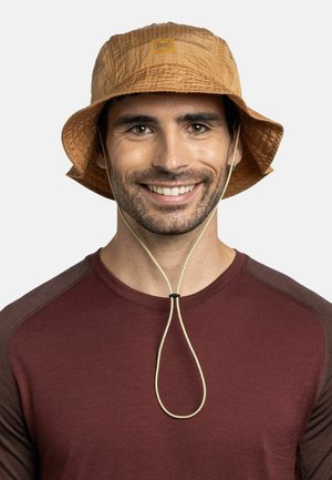 Hombre sonriente con sombrero de pescador marrón con correa en la barbilla y camisa de manga larga color granate, mirando hacia adelante sobre un fondo claro y liso.