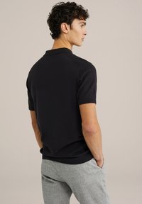 Polo shirt negru cu mâneci scurte, cu o textură netedă și guler ribat; prezentat pe un model din spate, asortat cu pantaloni gri deschis.