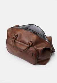 Sac de voyage en cuir marron avec deux poignées supérieures et poches zippées à l'avant. L'intérieur présente une doublure rayée. Surface douce et texturée.