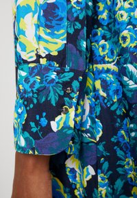 Blouse florale avec une base bleu marine foncé ornée de fleurs bleu vif, vert et jaune, avec des détails de poignets boutonnés et une texture de tissu douce.