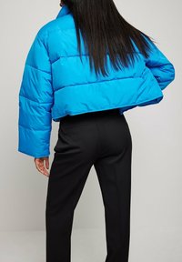 Chaqueta acolchada corta en azul brillante, con un diseño acolchado, mangas anchas y un acabado liso y brillante. Usada con pantalones negros.