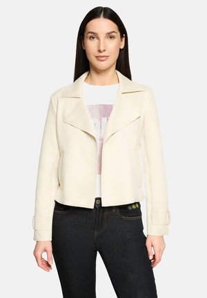 Veste courte couleur crème avec un large col, poches latérales et manches longues avec poignets boutonnés. Portée sur un t-shirt blanc et un jean foncé.