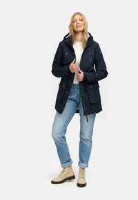 Námornícka parka bunda s kapucňou, zapínaním na zips a gombík, viaceré predné vrecká, nastaviteľný pás a pohodlné nosenie cez biely top.