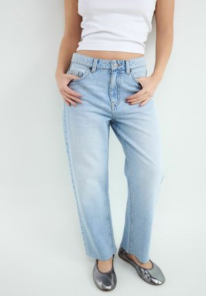 Žena nosi svetlomodré rifle s vysokým pásom, biely krátky top a metalické strieborné baleríny, stojí s rukami v vreckách.