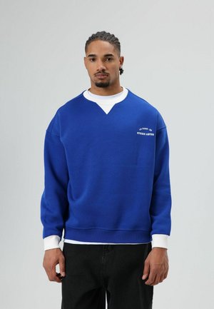 Blauwe sweater met een witte ronde hals, geribbelde mouwen en een V-hals ontwerp. Voorzien van het "STUDIO ABYSS" logo in het wit op de borst.